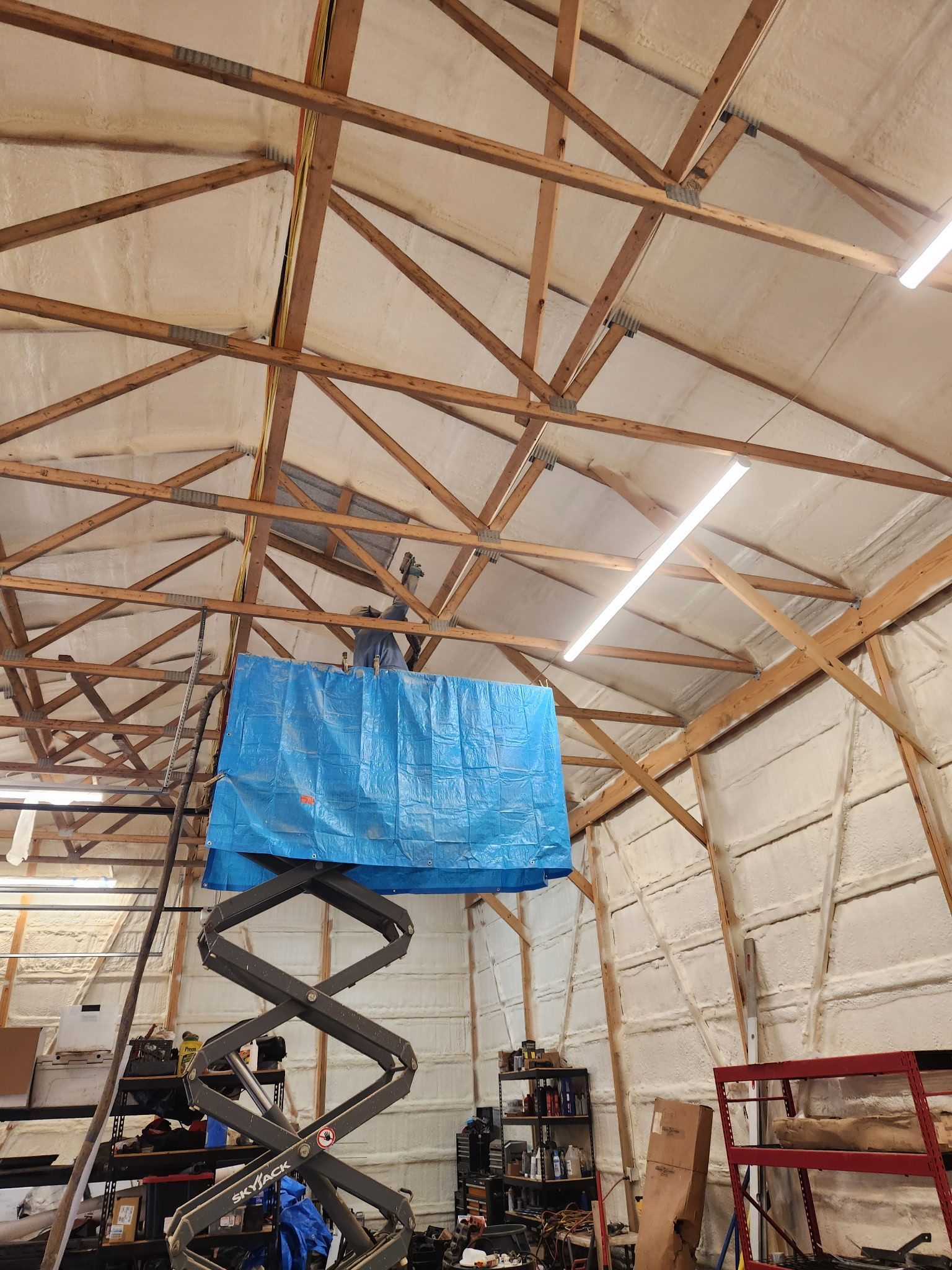 Person on a scissor lift working inside a wooden-framed building, with blue tarp and overhead lighting.