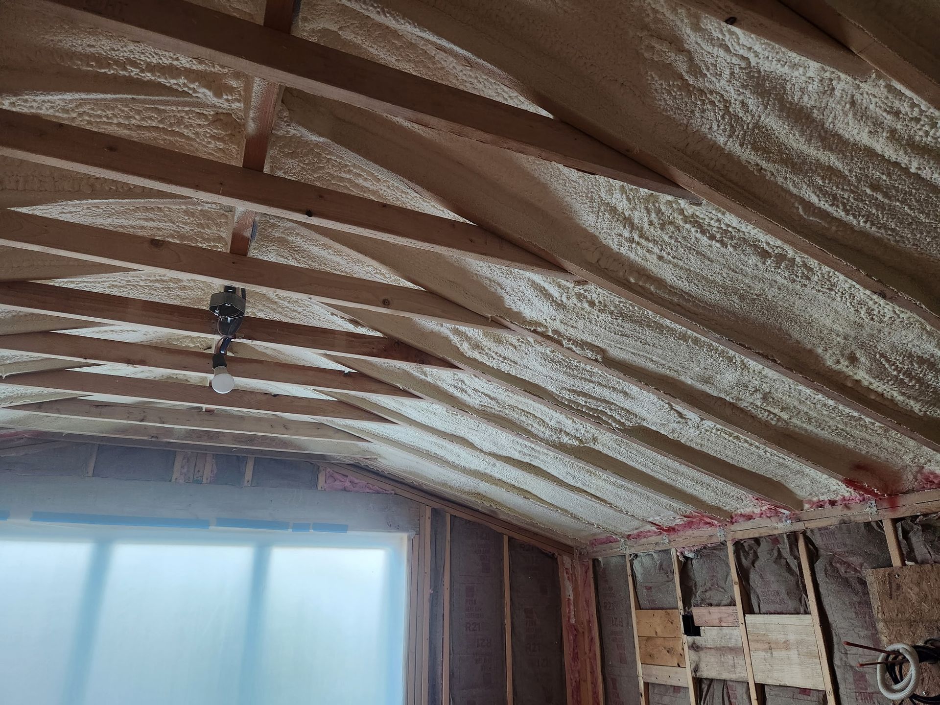 Interior view of an attic with spray foam insulation on the rafters and walls, a window visible on the left.