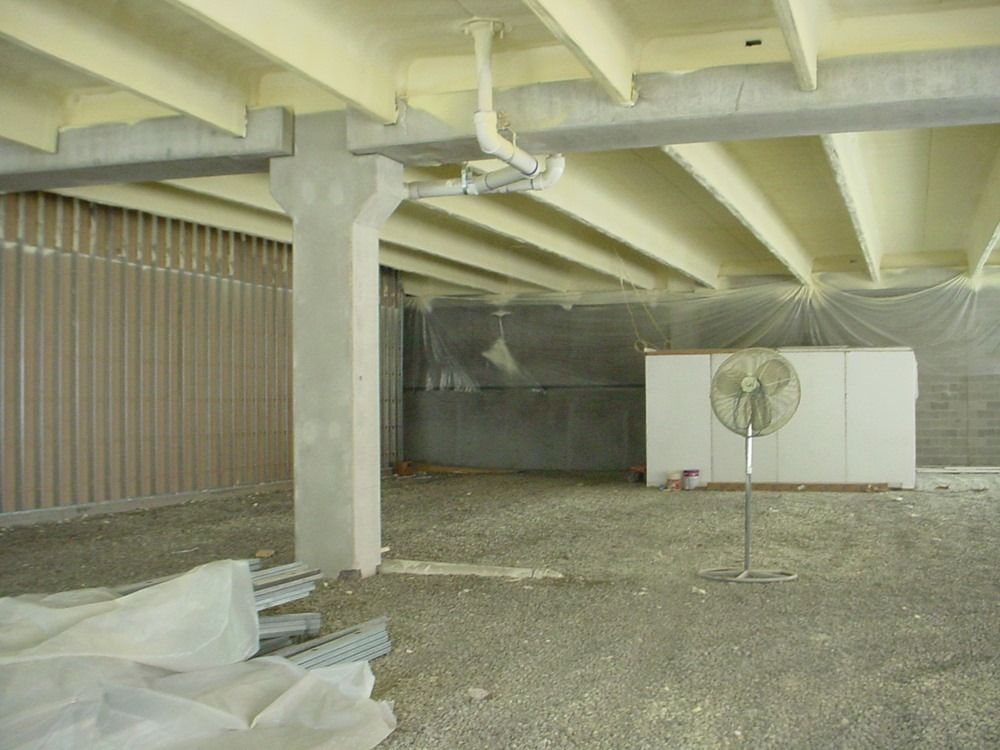 Interior of a building under construction, exposed beams, spray foam insulation, concrete floor, and a fan.