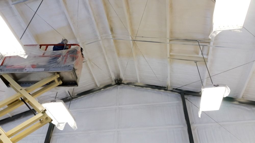 Person on a lift working on the white ceiling of a large warehouse; exposed metal beams, bright lights.