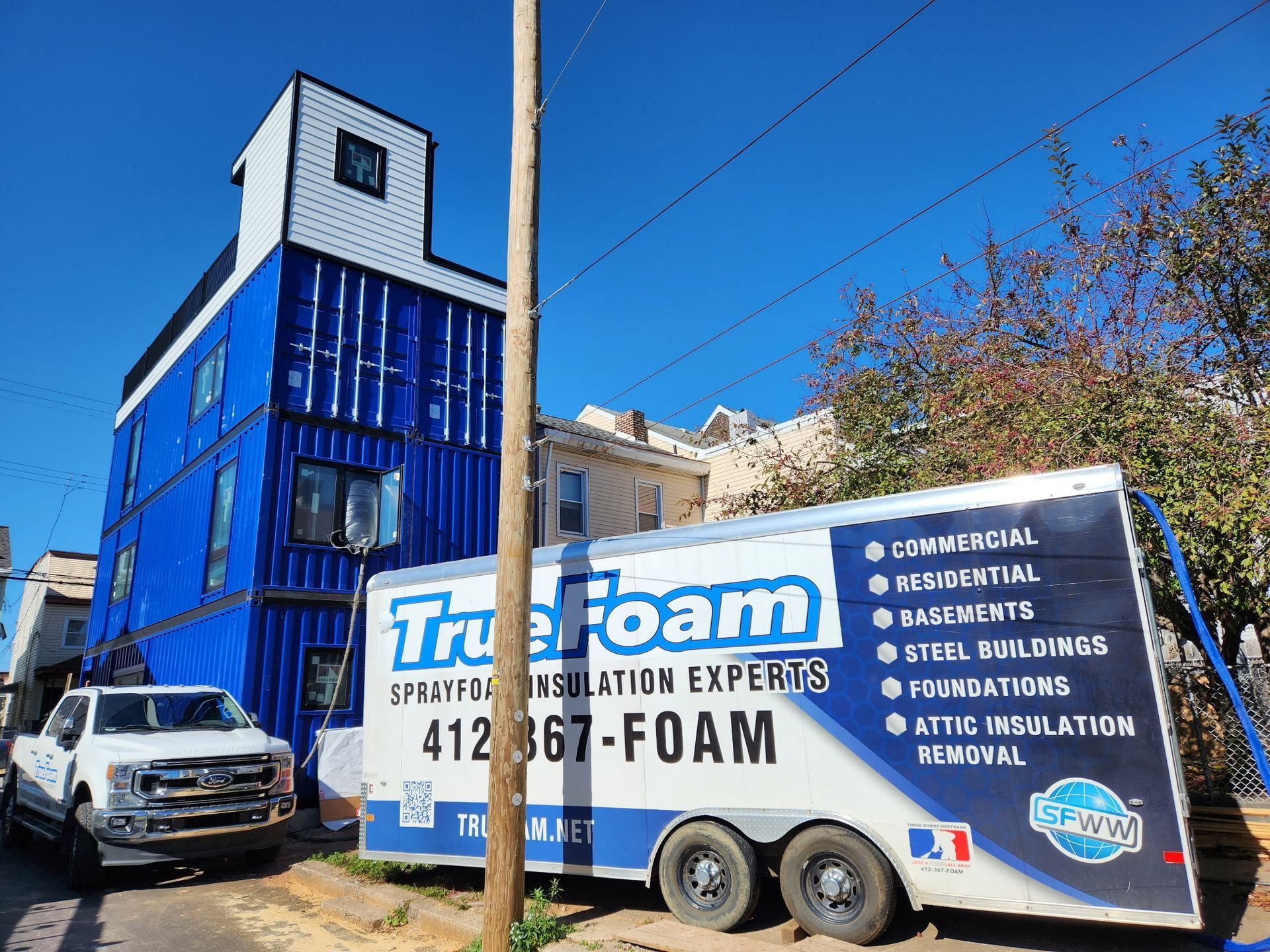 Blue shipping container home with True Foam trailer and truck, against a blue sky.