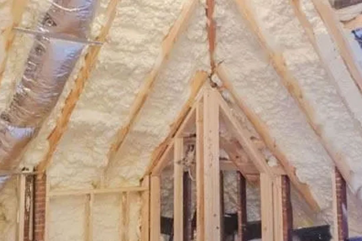Attic with exposed wood framing, spray foam insulation filling the spaces, and silver ductwork.