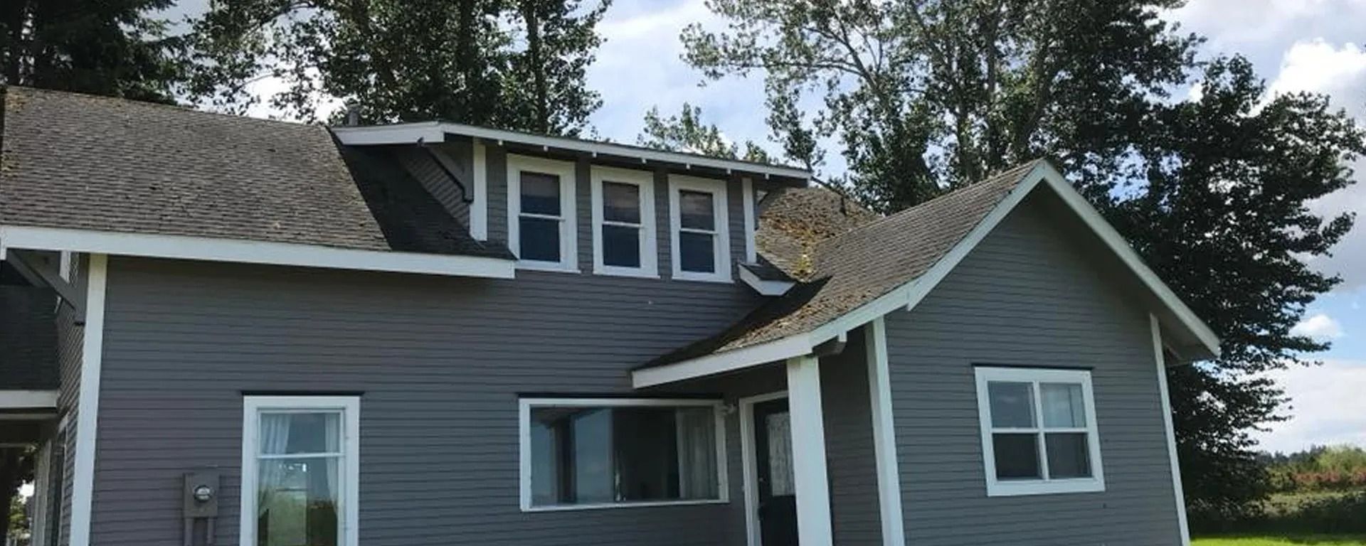 Gray house with white trim and dormer windows, seen from the side among trees and grass