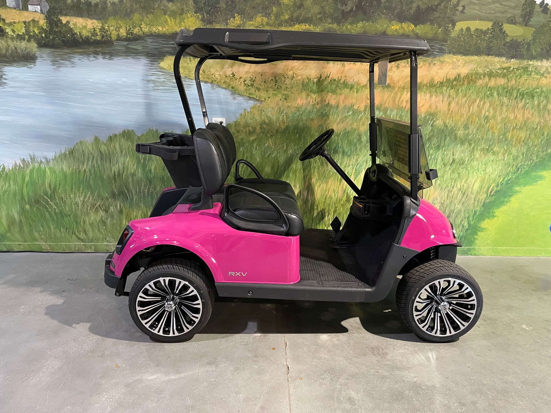 Pink golf cart with black accents and wheels, parked indoors, with a painted landscape background.