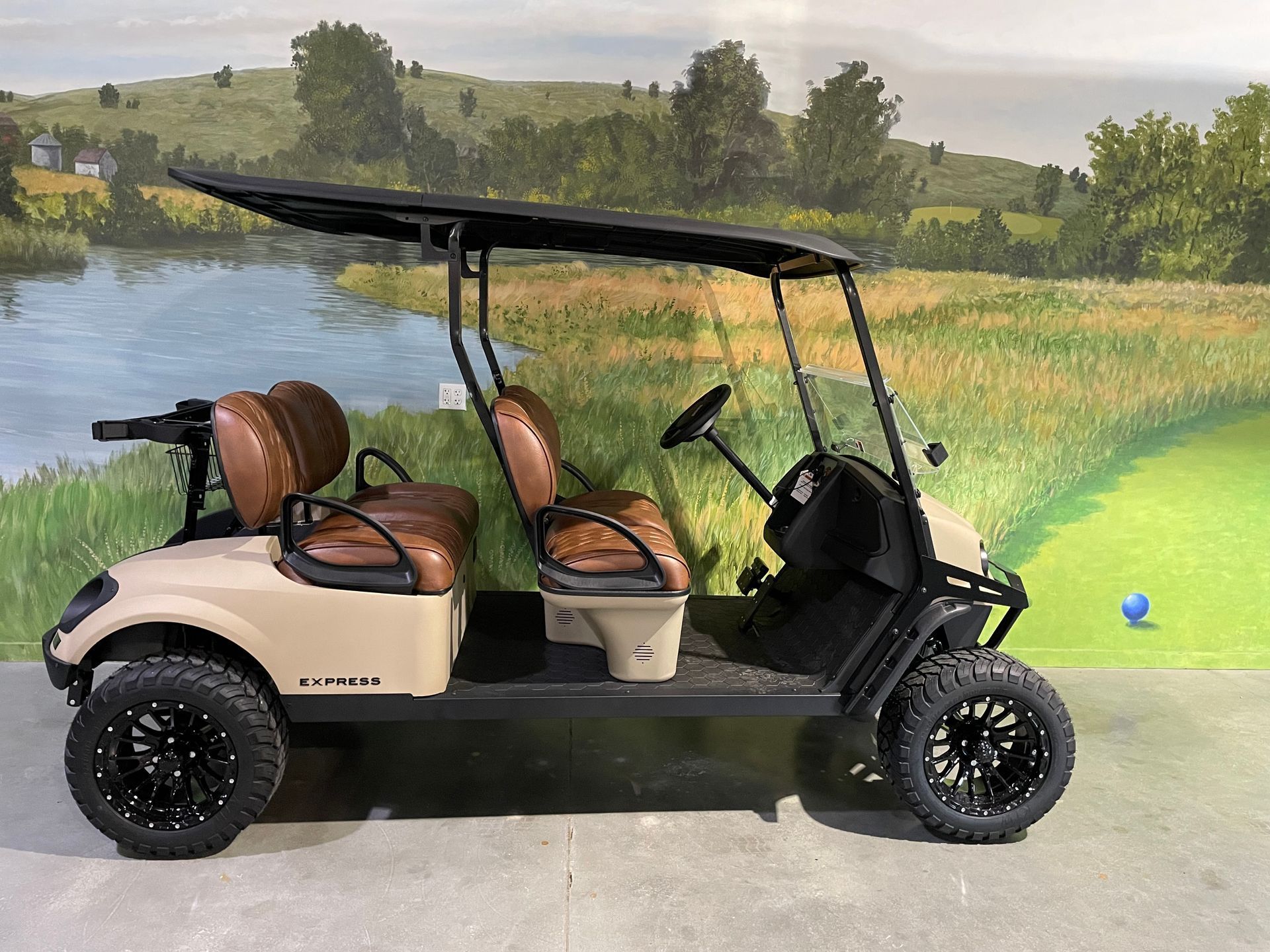 Tan golf cart with brown seats, black wheels, and a black roof, parked near a golf course.