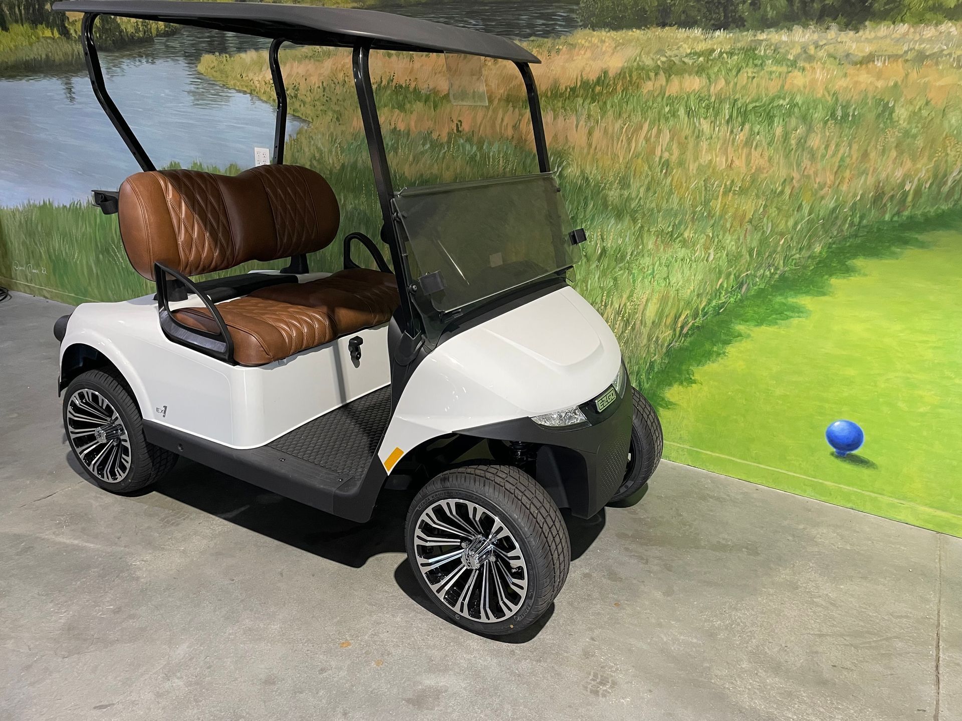 White golf cart with brown seats parked on a concrete surface, with a golf course background.