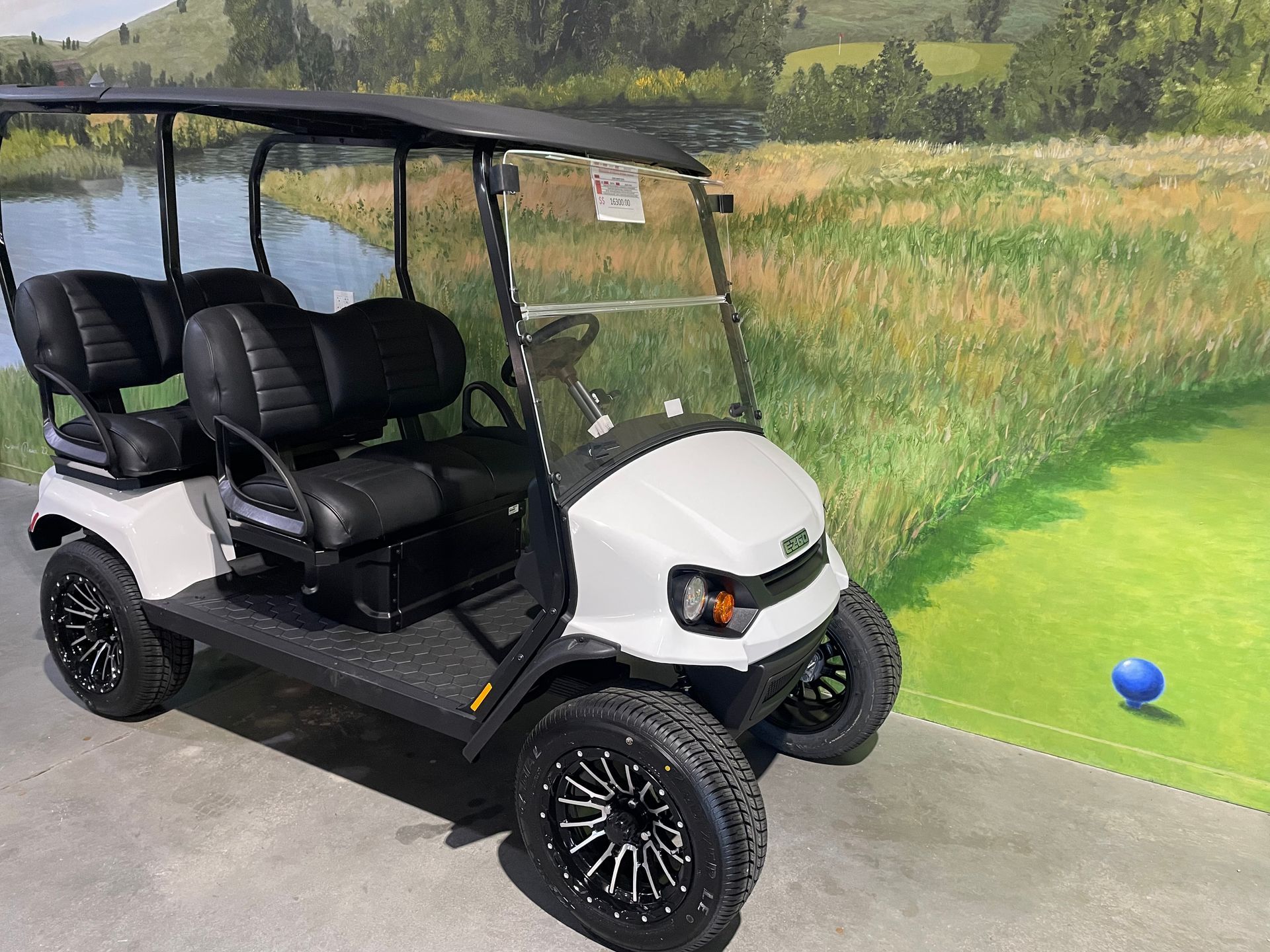 White golf cart with black seats and wheels, parked on a cement surface with a golf course backdrop.