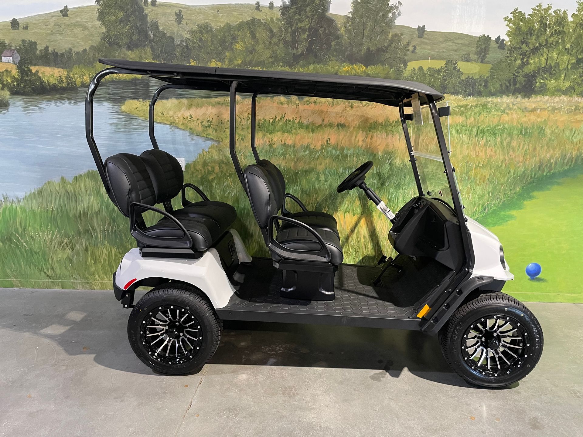 White golf cart with black seats and wheels, parked on a concrete surface in front of a painted landscape.