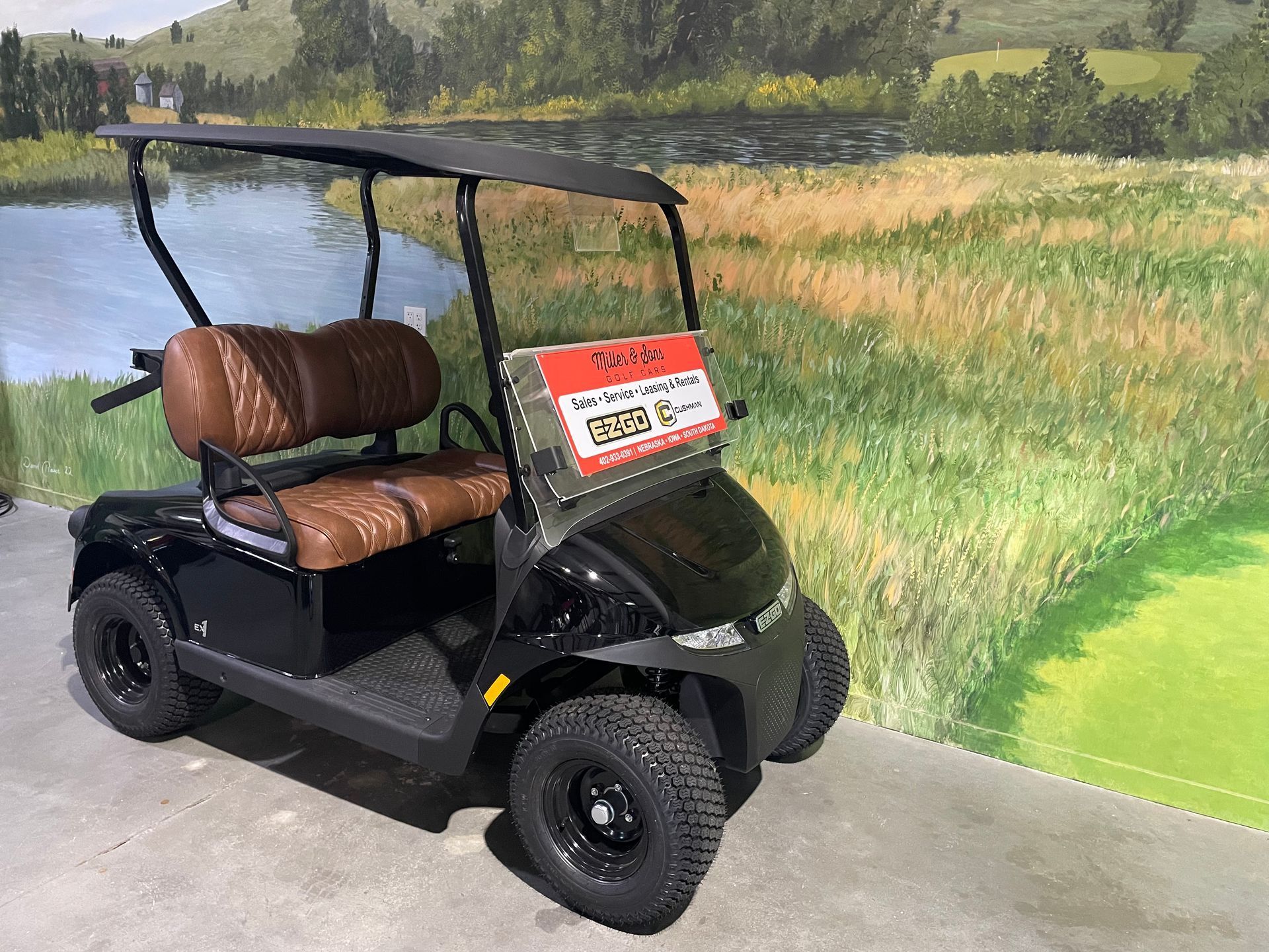Black golf cart with brown seats, parked indoors near a painted golf course backdrop.