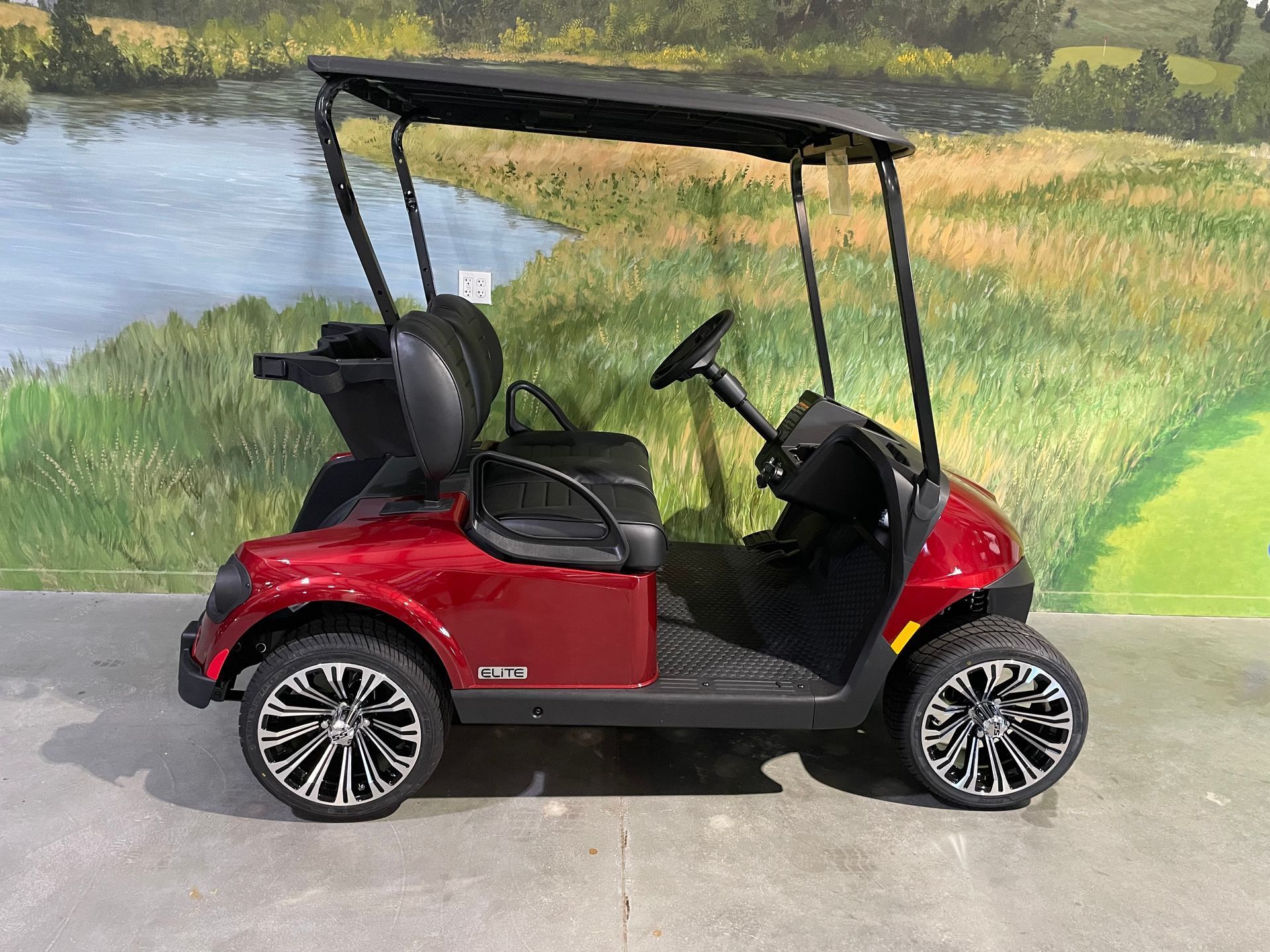 Red golf cart with a black roof and wheels, parked in front of a painted landscape.