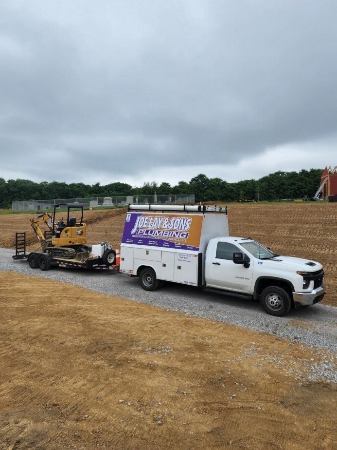 White truck with trailer hauling a mini excavator on a gravel road in a field under a cloudy sky.