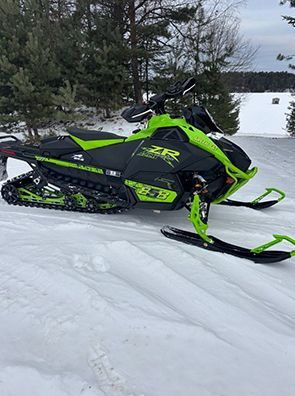 Green and black snowmobile parked in snow, near trees and a lake.
