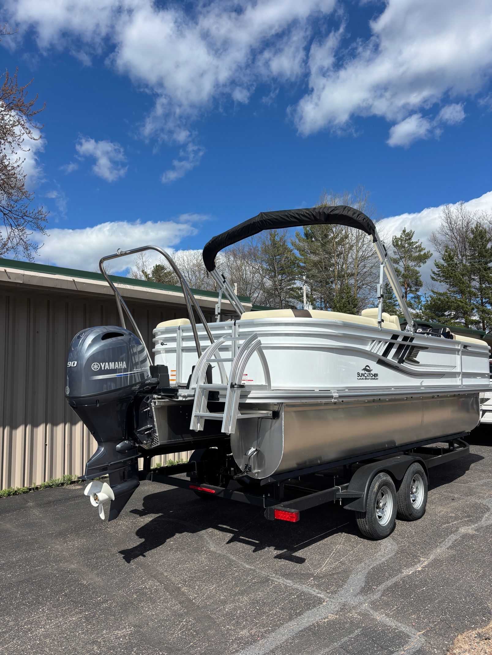 Pontoon boat on trailer with outboard motor parked beside a building under a blue, cloudy sky