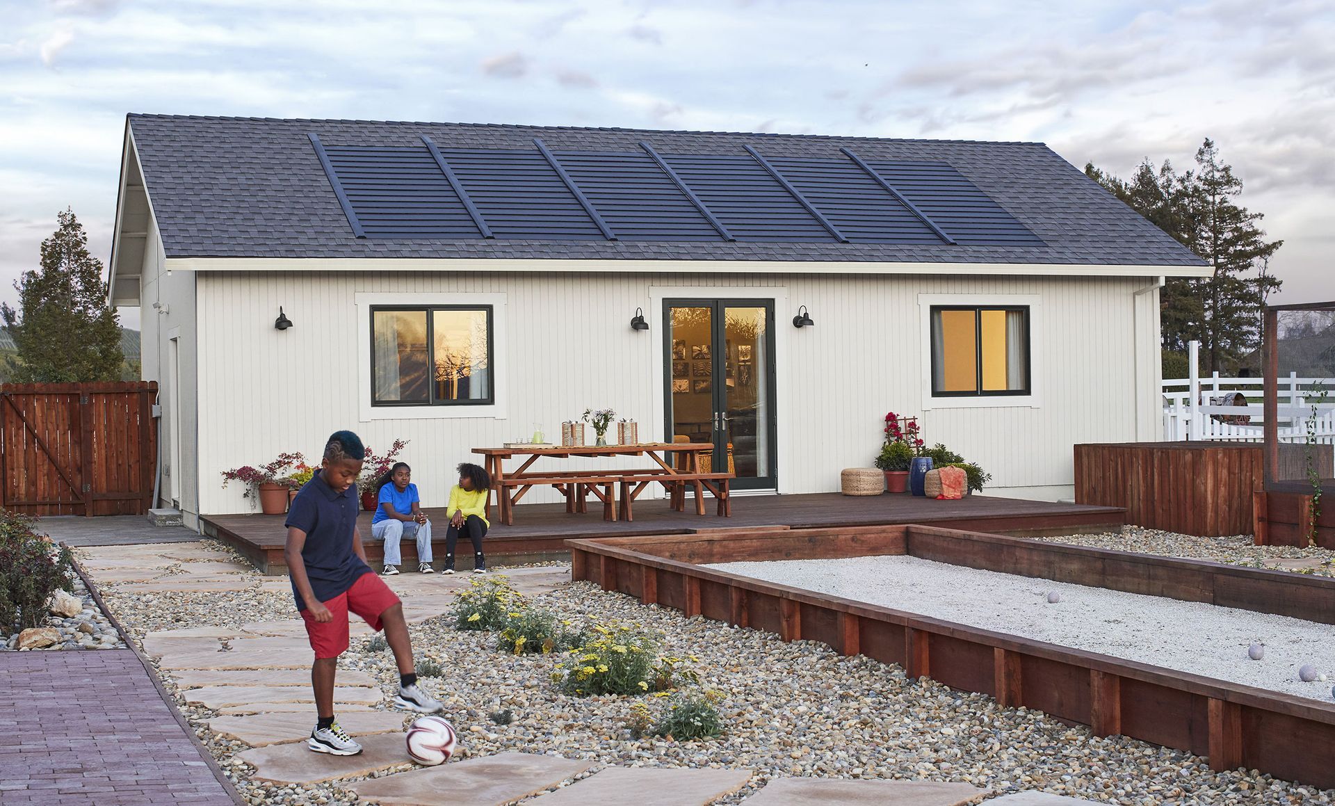 A boy is kicking a soccer ball in front of a house with solar panels on the roof.