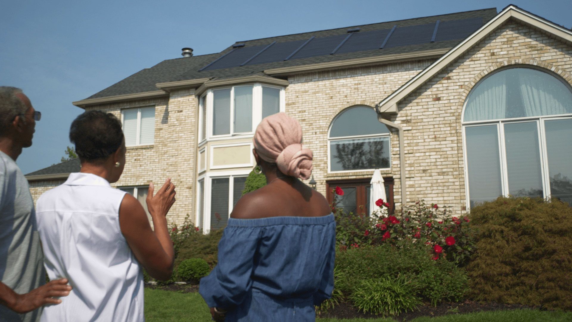 A group of people are standing in front of a large brick house with solar panels on the roof.