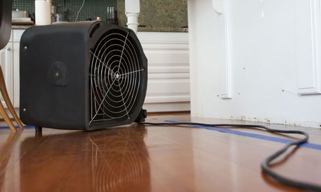 A black fan is sitting on a wooden floor in a kitchen.