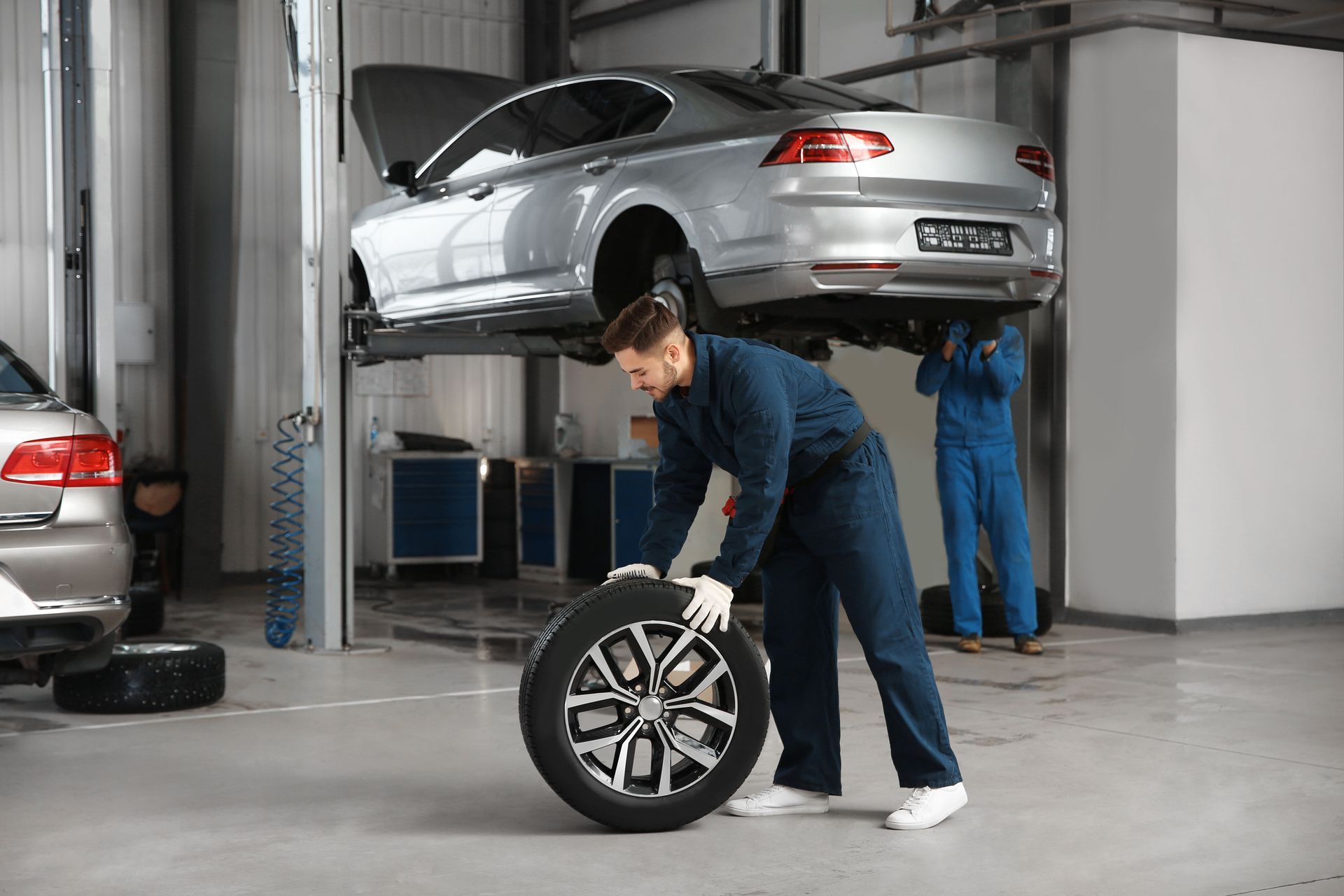 Mechanic in a garage lifting a car wheel, with another car raised on a lift behind him