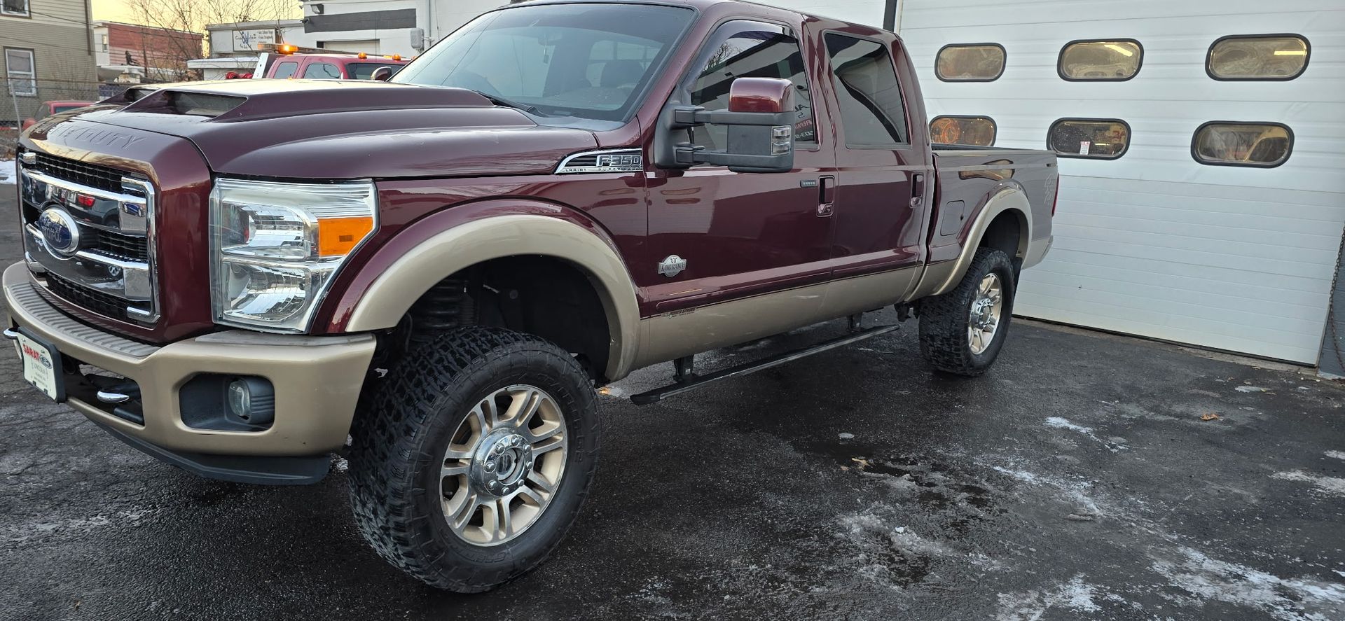 Maroon and tan lifted Ford pickup truck parked on a wet lot by a garage door