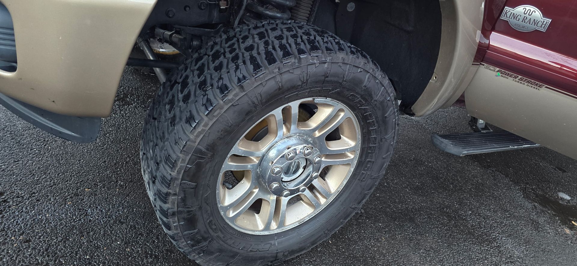 Close-up of a lifted tan and maroon truck’s chrome wheel and tire on wet pavement