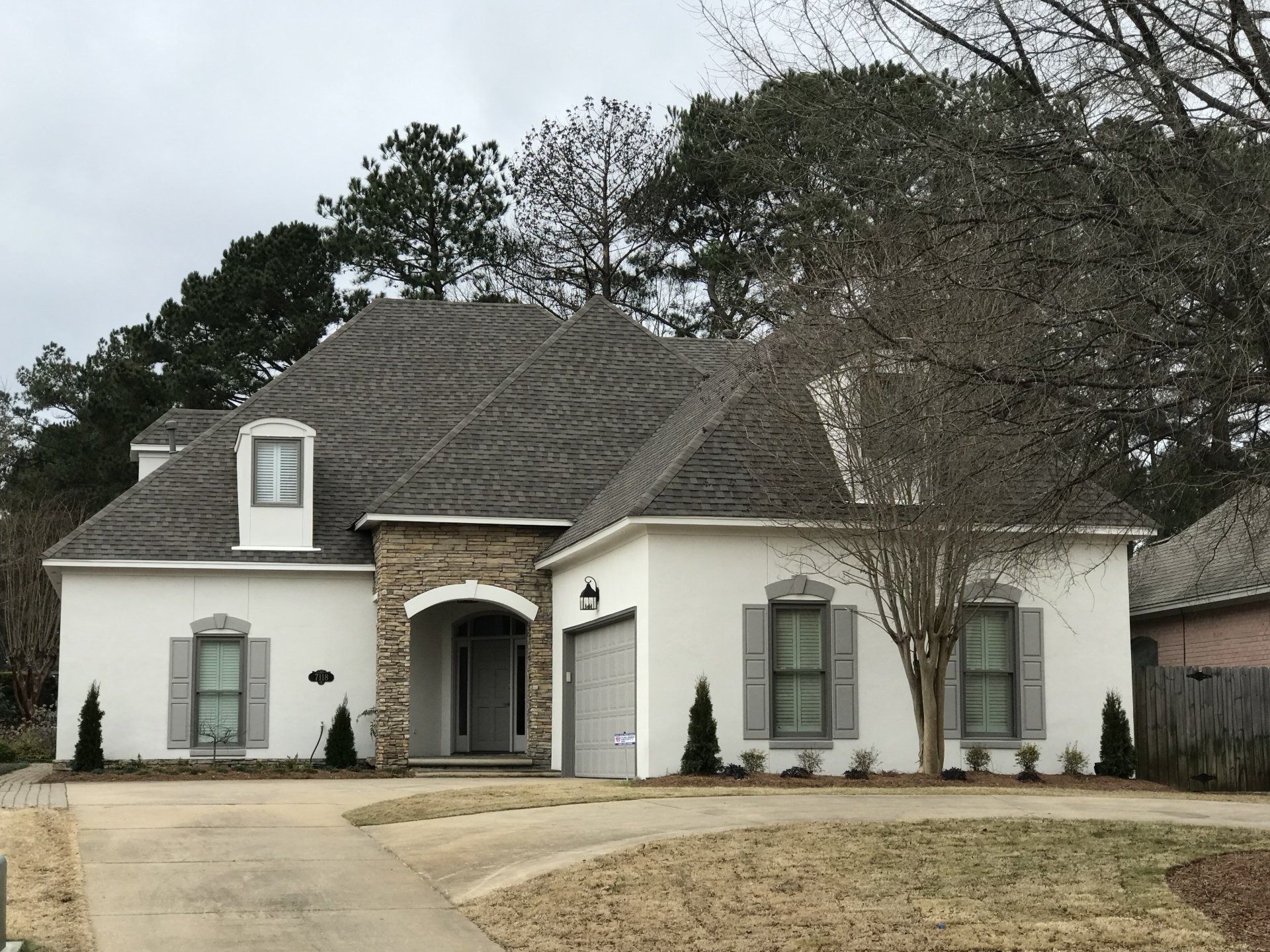 White house with gray roof, stone entryway, and shutters; trees in the background, overcast sky.