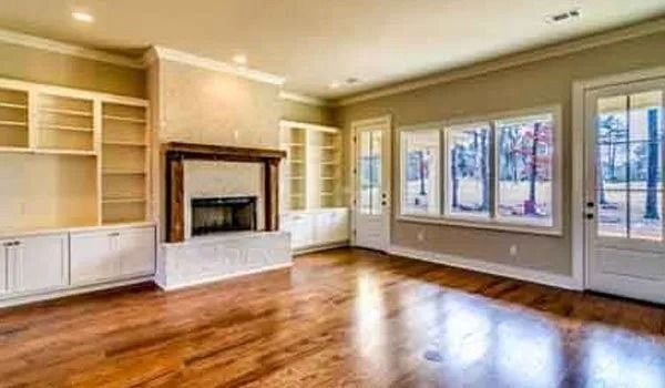 Empty living room with fireplace, built-in shelves, hardwood floors, and large windows.