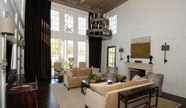 High-ceiling living room with large windows, dark curtains, light sofas, fireplace, and chandelier.