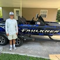 Man standing on a boat, wearing a long-sleeved shirt and shorts. Boat has “Faukner Bass Fishing” on the side.