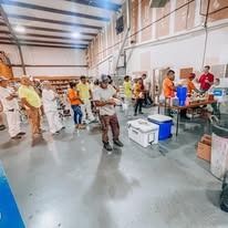 People in a warehouse setting, some at tables with food, others gathered near containers.