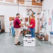 Two men in a warehouse; one holding a paint can. One wears a red shirt. Coolers are in front of them.