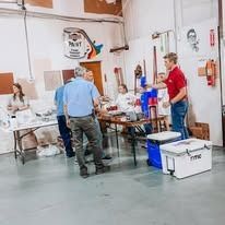 People in warehouse sorting supplies and equipment, including coolers, a paint sign, and tools.