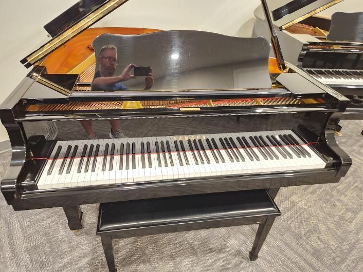 Black grand piano with open lid and keyboard, reflecting a person taking the photo.