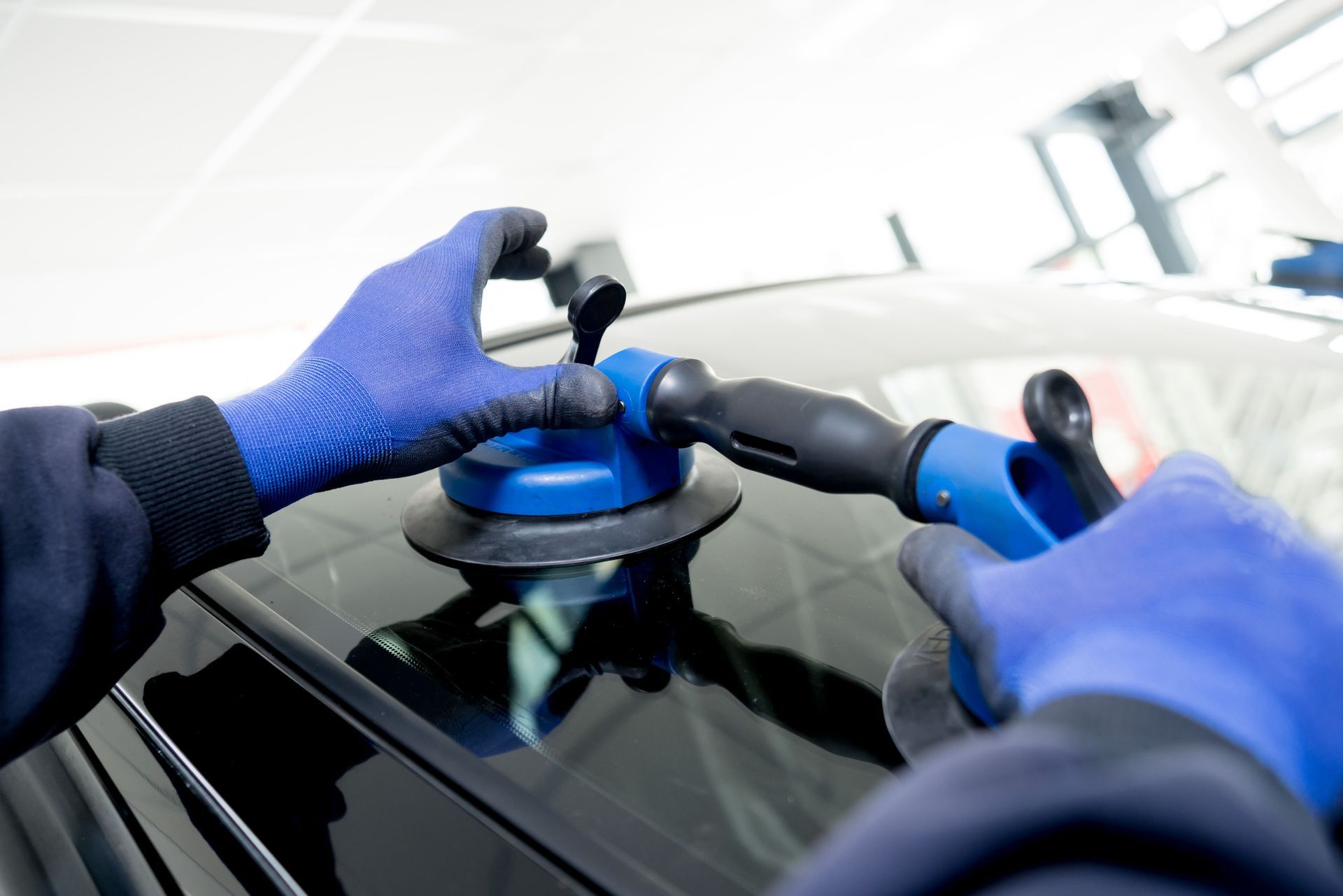 Gloved hands polishing a car windshield with a blue buffer in a bright auto shop