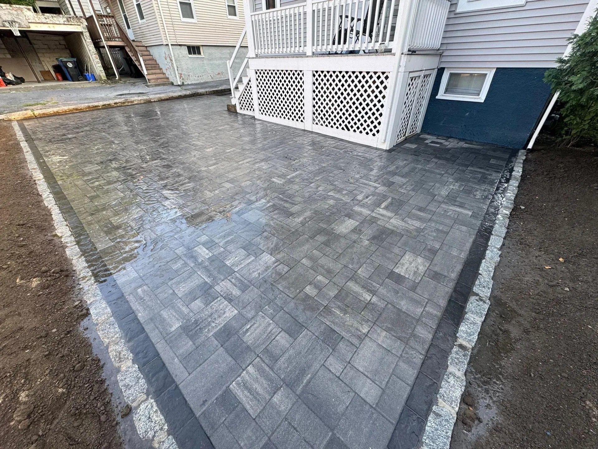 Wet gray paver driveway beside a house with white lattice porch and a dark border pattern.