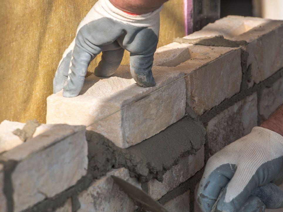 Gloved hands laying light-colored bricks on a wall with wet mortar