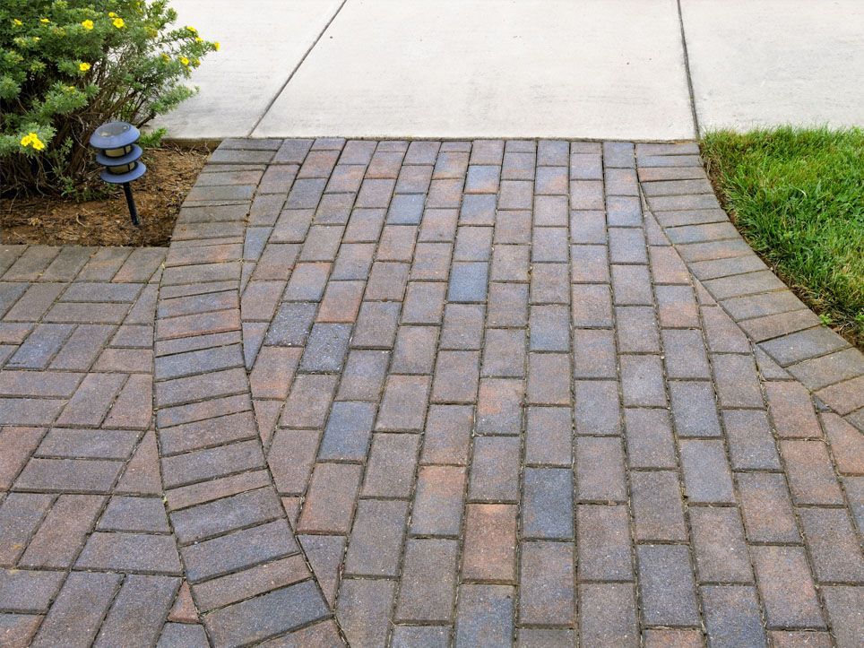 Brick walkway leading to a concrete driveway, bordered by grass and a small garden light