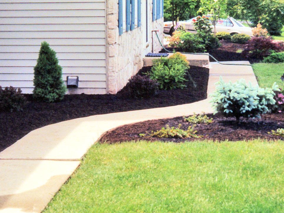 Curved concrete walkway beside a house with mulch beds, shrubs, and green lawn