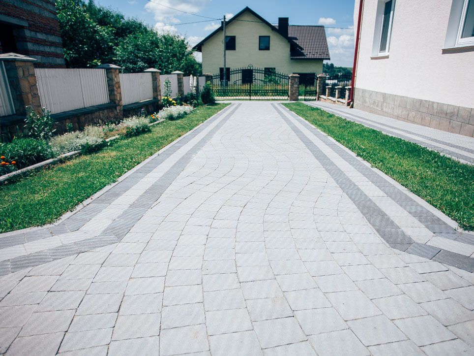 Paved driveway leading to a house, bordered by green grass and flower beds under a partly cloudy sky