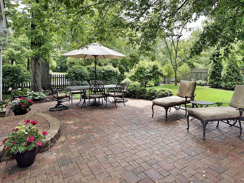 Brick patio garden with outdoor table, umbrella, lounge chairs, and potted flowers amid lush greenery