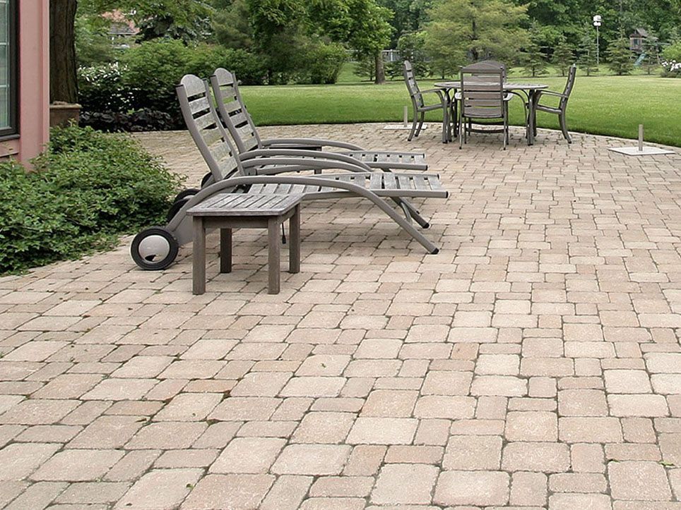 Patio with stacked lounge chairs, a table set, and a paved walkway beside green shrubs and trees.