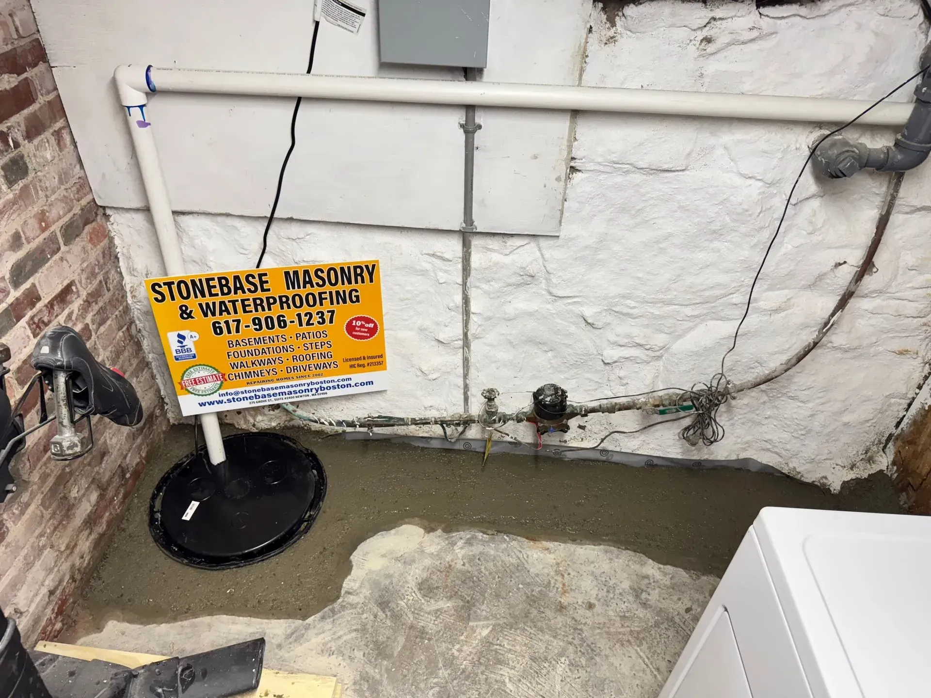 Sump pump installation in a basement with concrete and brick walls. Piping, electrical box, and business sign are also visible.