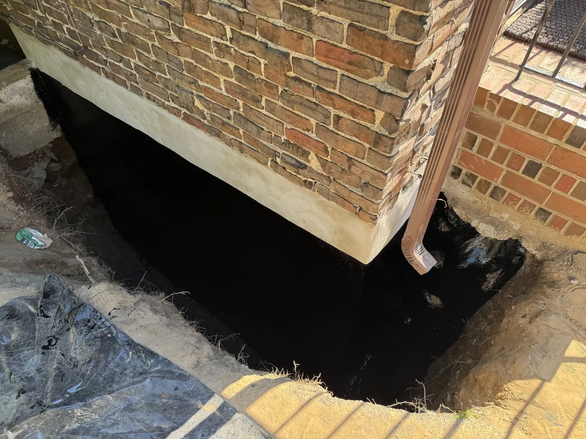 Black waterproofing layer in a trench next to a brick wall and a downspout.