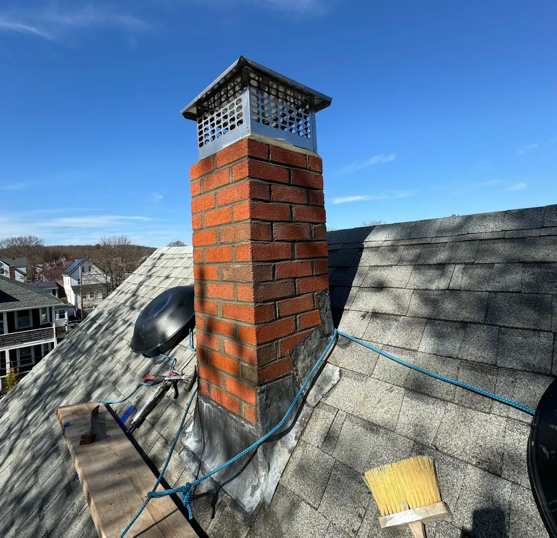 Brick chimney on a shingle roof with a metal cap, under a blue sky.