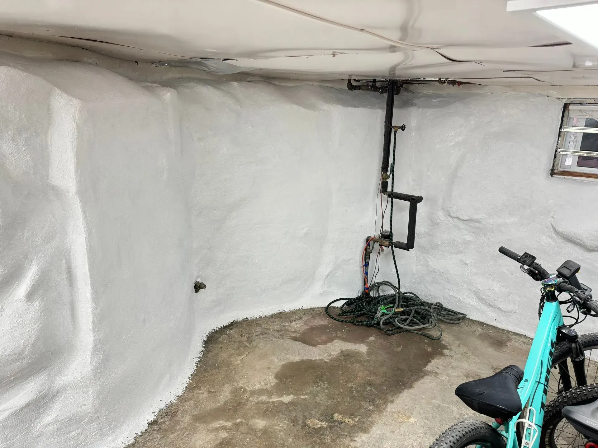 White spray foam insulation coats basement walls, with pipes and a bike present.