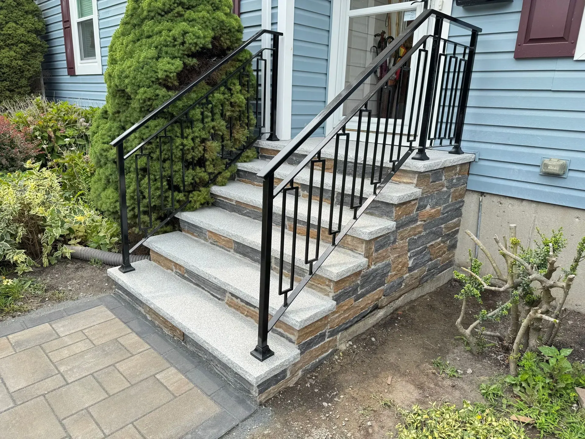 Black metal handrails on gray stone steps leading to a blue house with brick accents and landscaping.