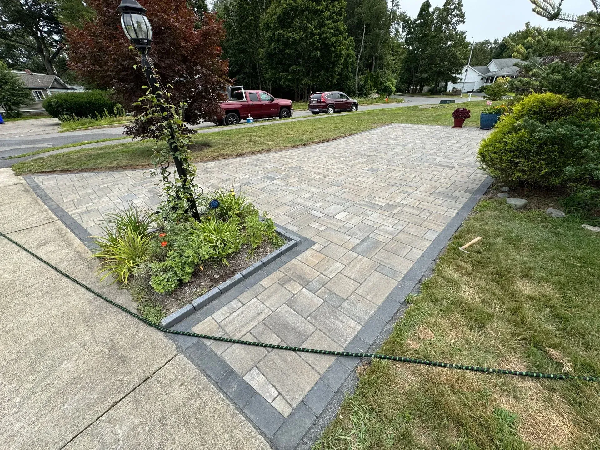 Brick paved driveway with a small garden bed and a street in the background. Cars are parked nearby.