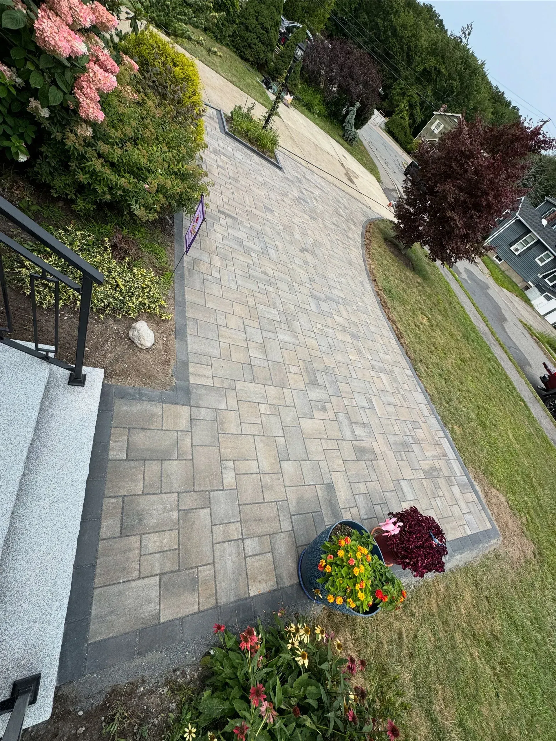 Brick paved driveway curving to the right, bordered by grass and flowerbeds.