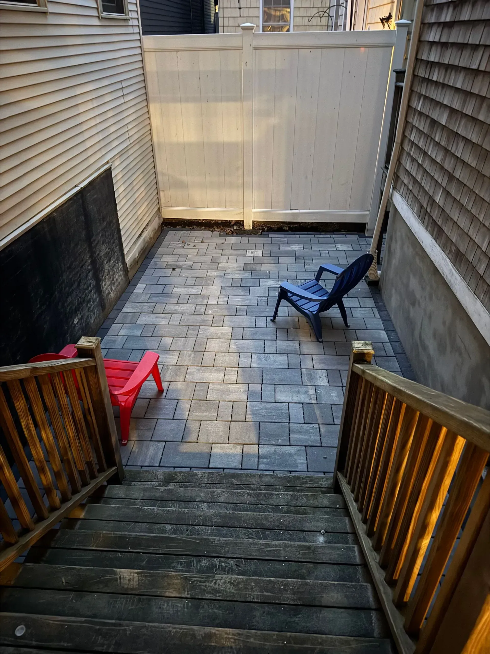 View from wooden stairs down to a small, paved backyard with two chairs and a white fence.
