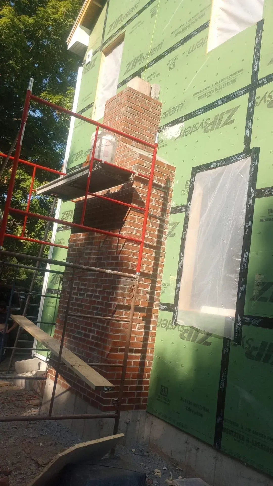 Brick chimney under construction on a house, with scaffolding and green sheathing visible.