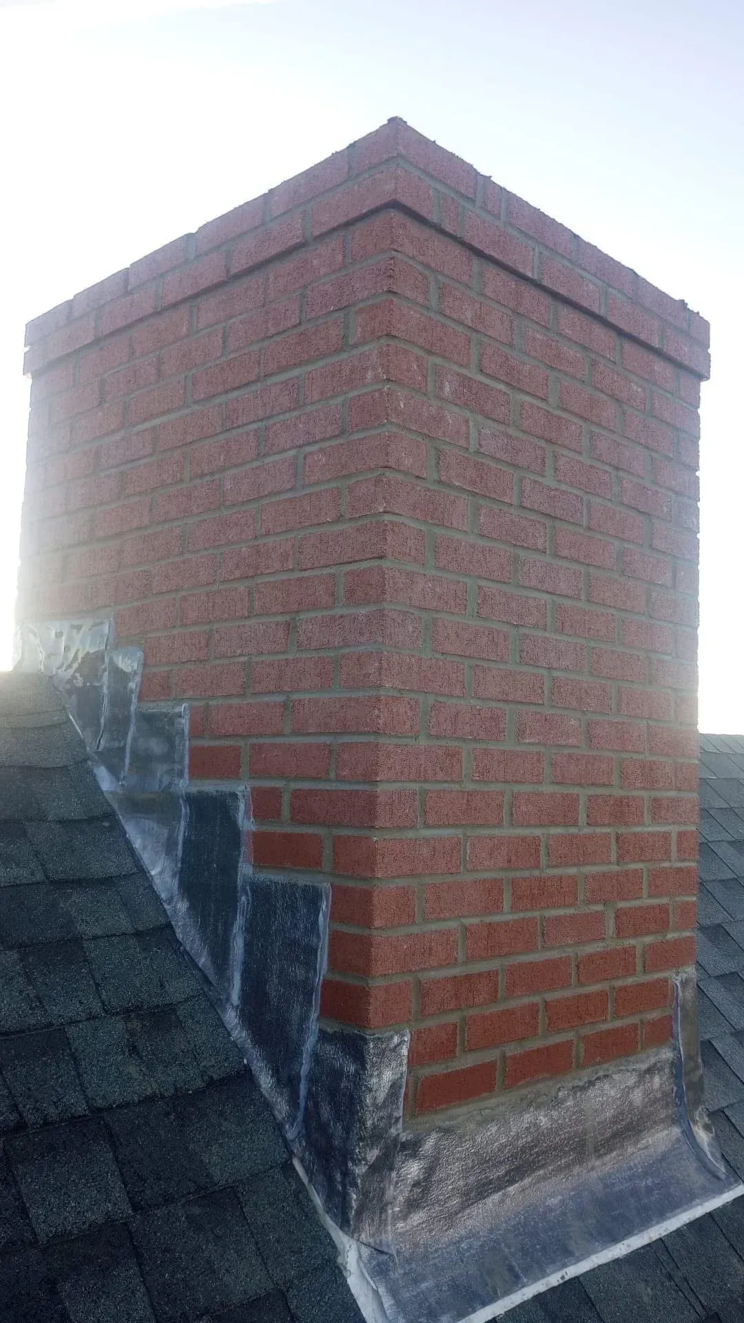 Brick chimney on a roof with gray shingles and metal flashing.