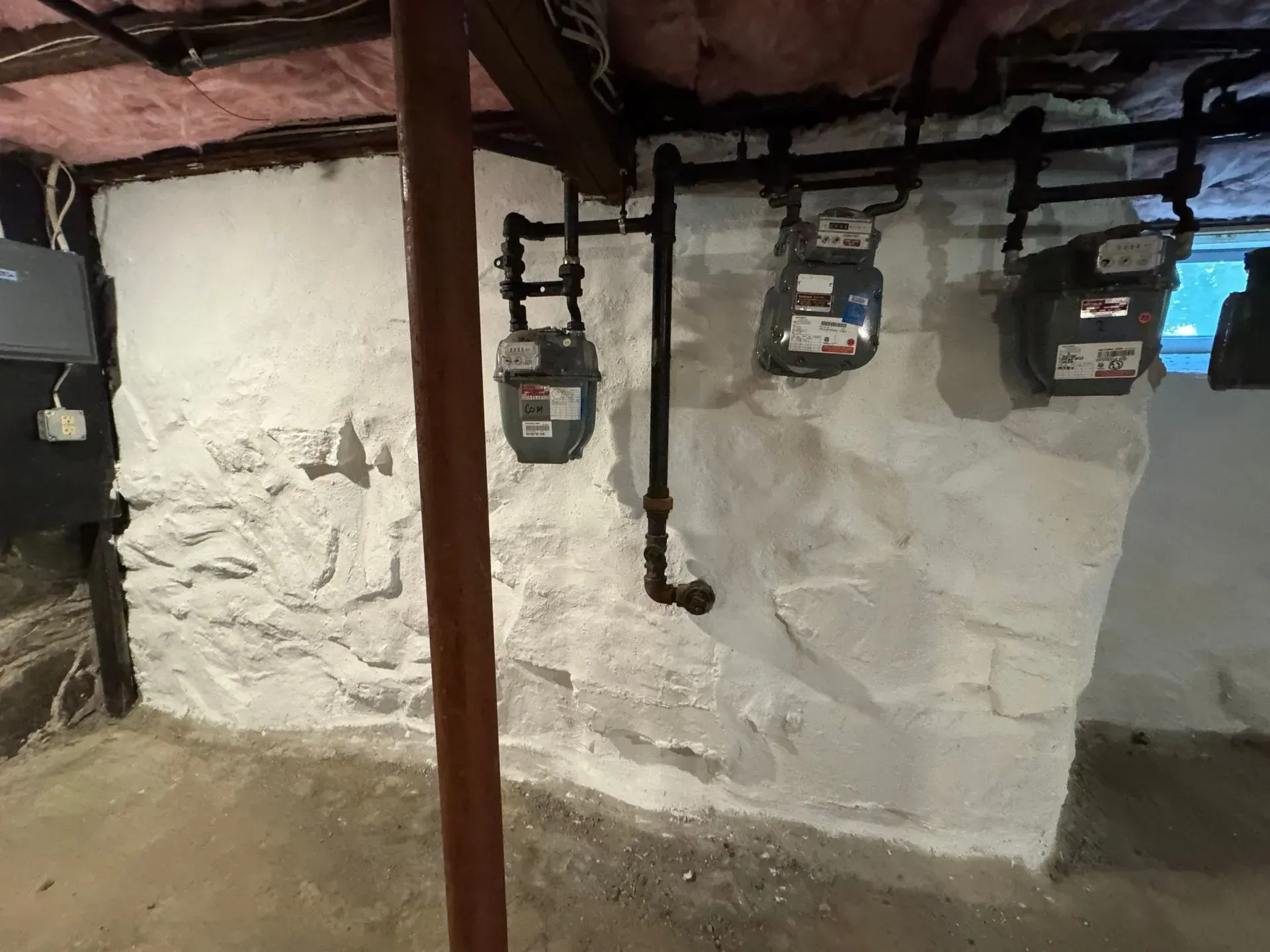 Basement with stone wall, gas meters, and pipes. The wall is painted white, with a dark pipe and wooden beam overhead.